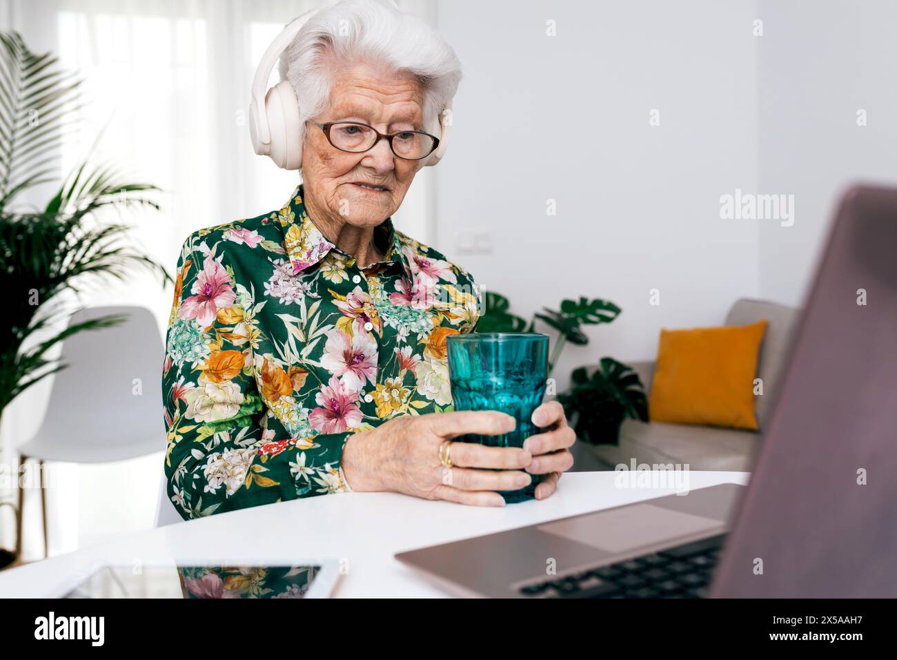 Elderly lady in a floral shirt enjoys a glass of water while working on her laptop in a bright living room, showcasing active aging and technology use Stock Photo
