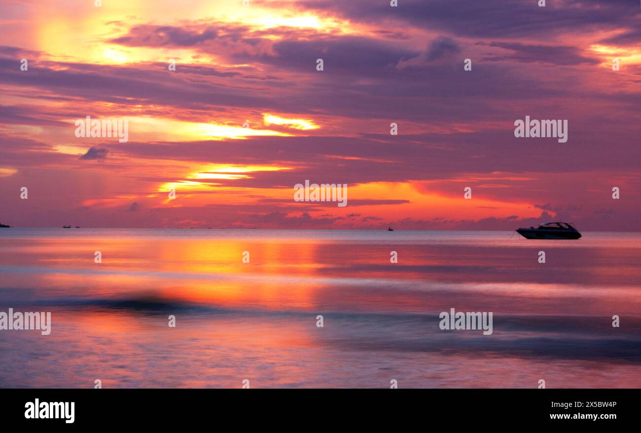Dawn at Chaweng Beach, Koh Samui, looking out at sea, the sun partly hidden by clouds, reflecting off the water, with the boat a silhouette. Stock Photo