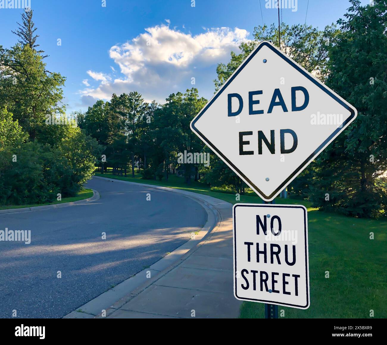 White Warning Road Signs on DEAD END street and NO THRU STREET along road and sidewalk in evening. Stock Photo