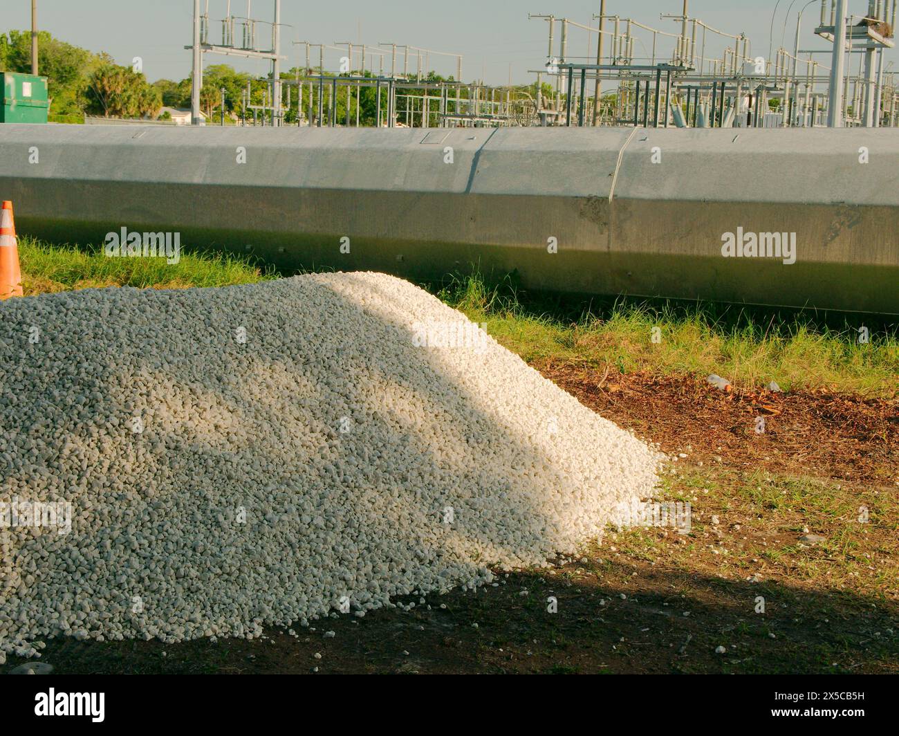 Wide view over green grass to pile of white stones and laying on the ground a steel Utility Pole sections with a Electric Substation in back. Palm Stock Photo