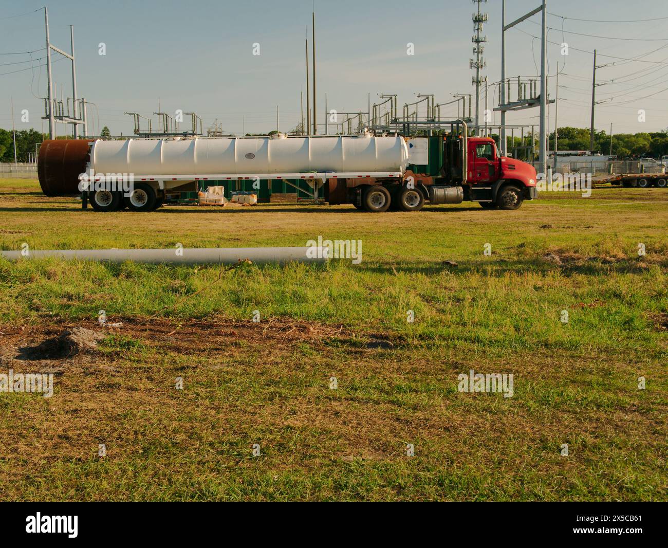 Wide view over green grass to a steel Utility Pole section laying ground a with a water truck and Electric Substation in back. afternoon. Blue sky. Stock Photo