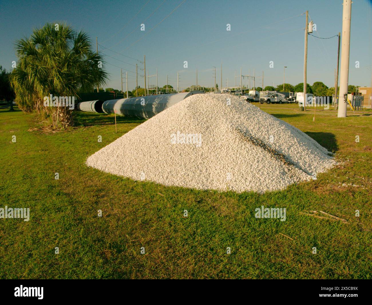 Wide view over green grass to pile of white stones and laying on the ground a steel Utility Pole sections with a Electric Substation in back. Palm Stock Photo