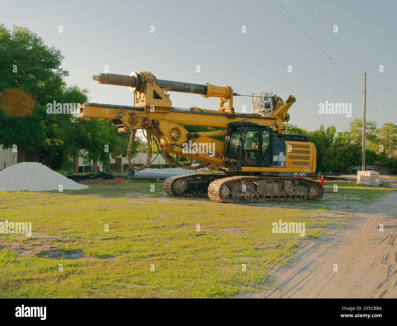 Wide view of a large yellow ROTARY DRILLING RIG in grass and dirt with a pile of small white stones on the left and steel utility poles sections. Stock Photo