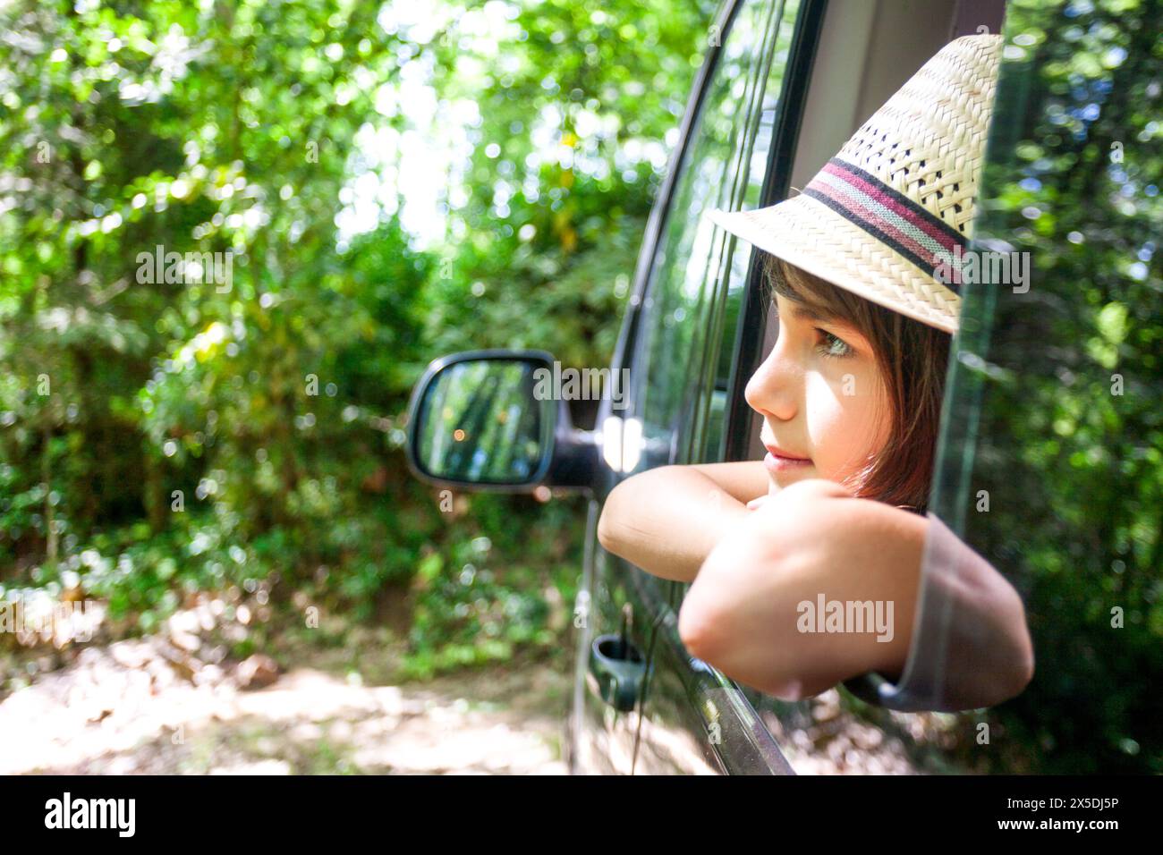 A young boy is looking out the window of a car. He is wearing a straw hat and has her arm out the window. The scene is peaceful and relaxing, as the b Stock Photo