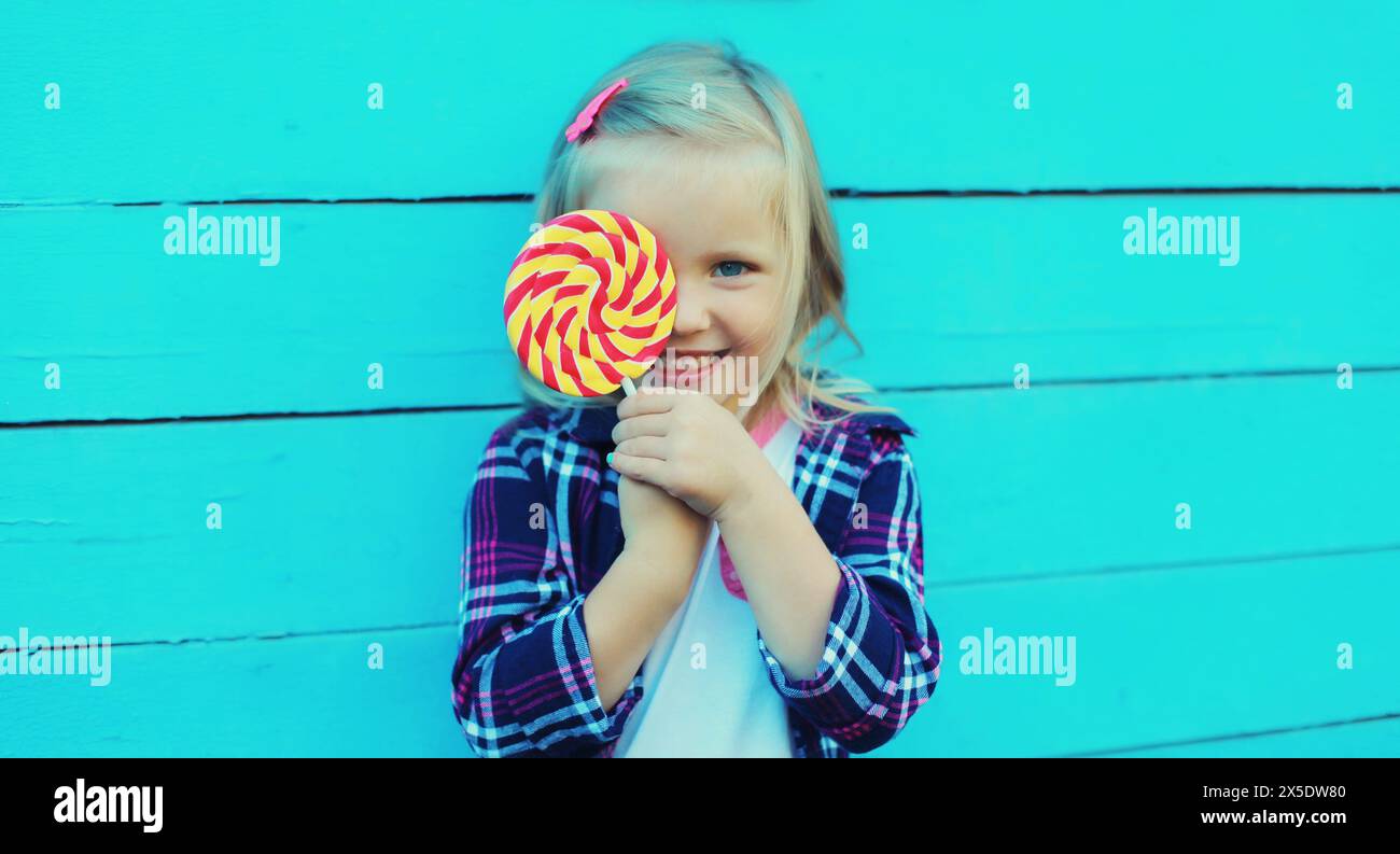 Portrait of happy cheerful smiling little girl child with sweet lollipop on stick on blue background Stock Photo