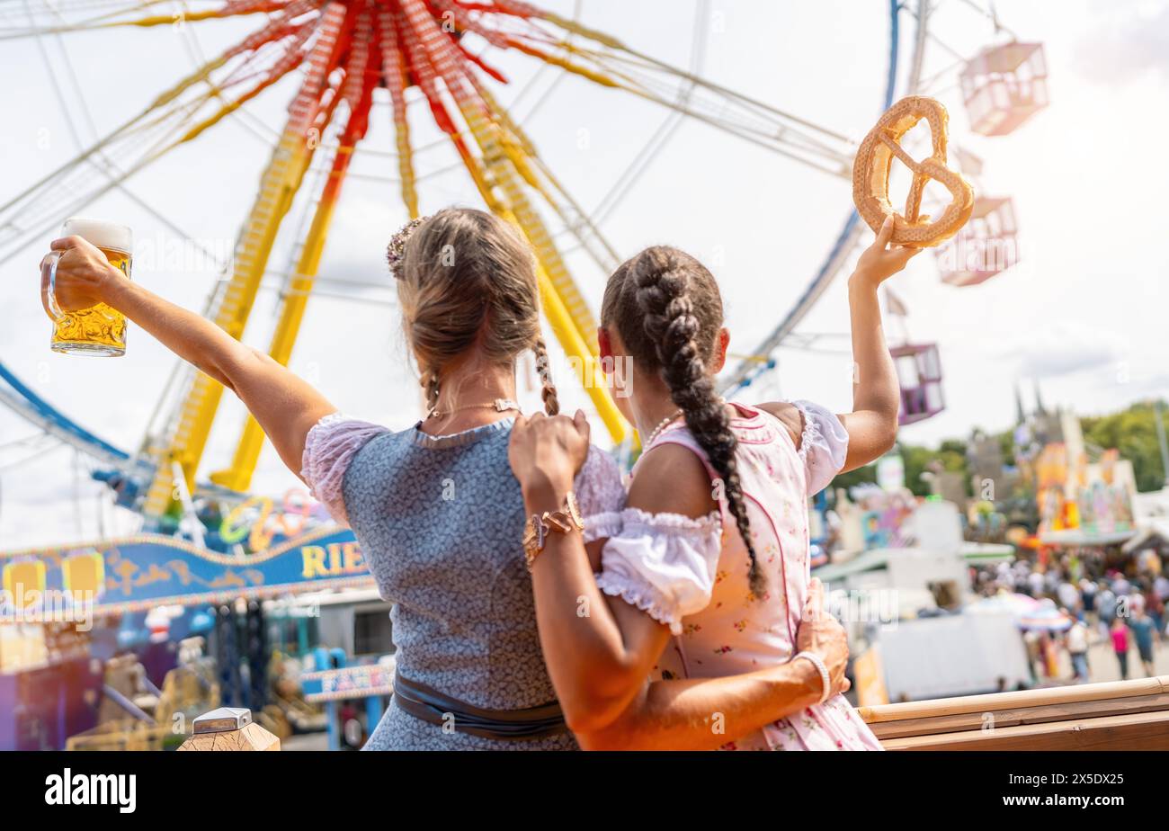 bavarian women in tracht holding a beer mug and pretzel aloft at a lively fairground with Ferris wheel at oktoberfest or duld in germany Stock Photo