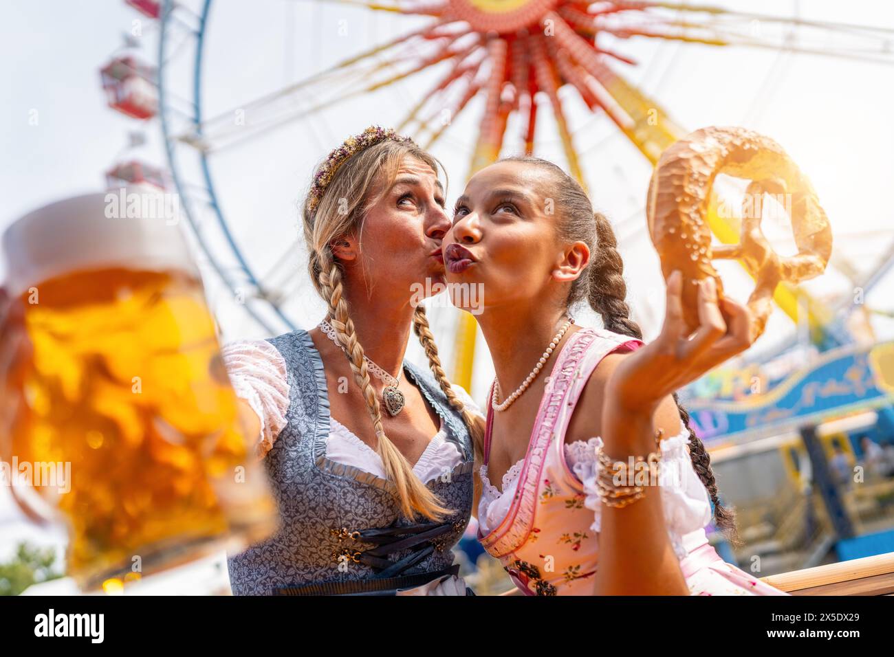 Girlfriends in a traditional Bavarian dress tracht gives her friend a kiss on the cheek, holding beer and a pretzel at oktoberfest or duld in germany Stock Photo