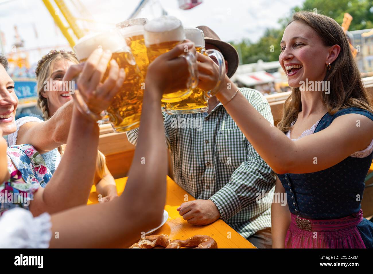 Joyful friends clinking beer mugs at a oktoberfest or dult festival beer tent table, amusement rides and blue sky in the background Stock Photo