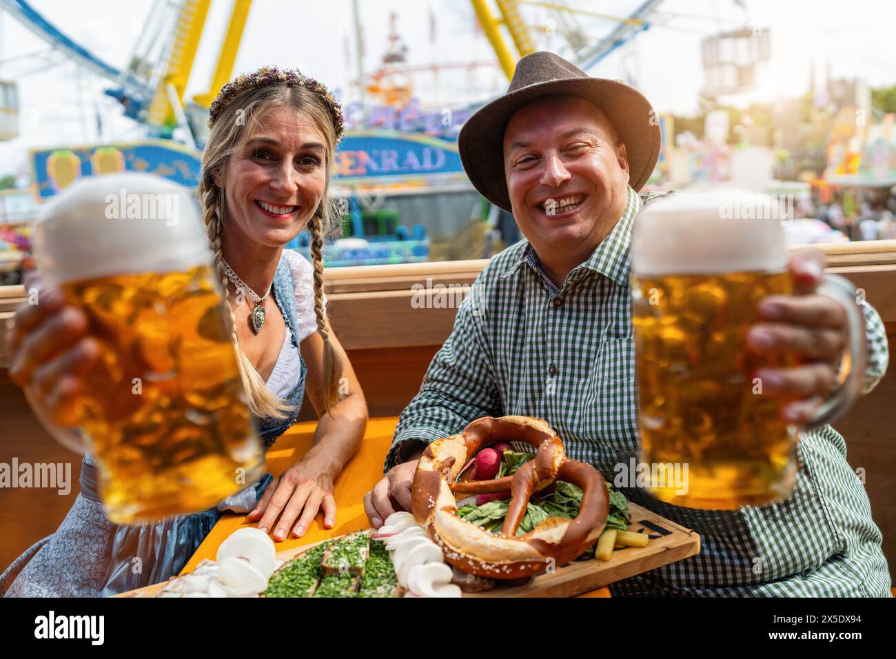 Man and woman in traditional outfits raising beer mugs at a oktoberfest festival, with food on the table and rides in background in munich germany Stock Photo