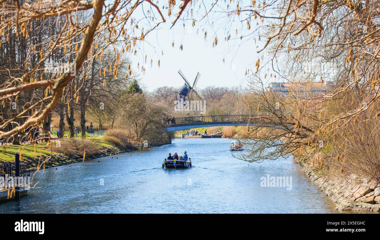 Leisurely boat tour along a scenic canal framed by budding trees with a traditional windmill in the background, in the heart of Malmö. Concepts: relax Stock Photo