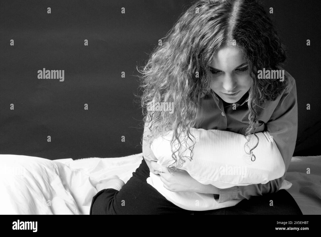 Young woman clutching a pillow to her stomach while sitting on bed, depicting the concept of stomachache or stomach cramps, USA. Stock Photo