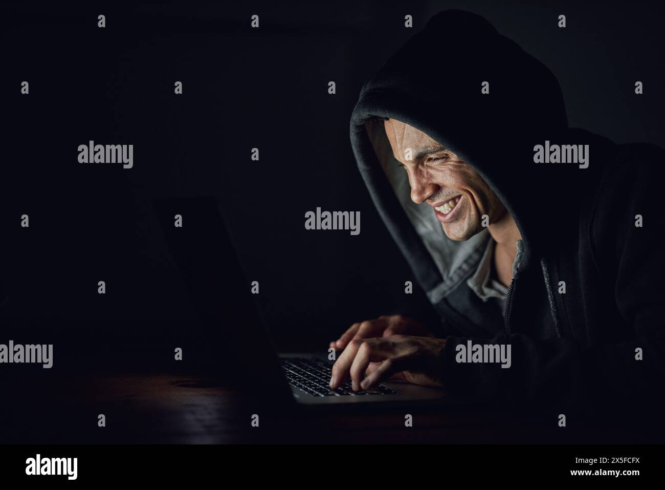 Dark, basement and man at laptop for hacking, online programming and cybersecurity at night. Software, developer or hacker in hoodie coding for Stock Photo