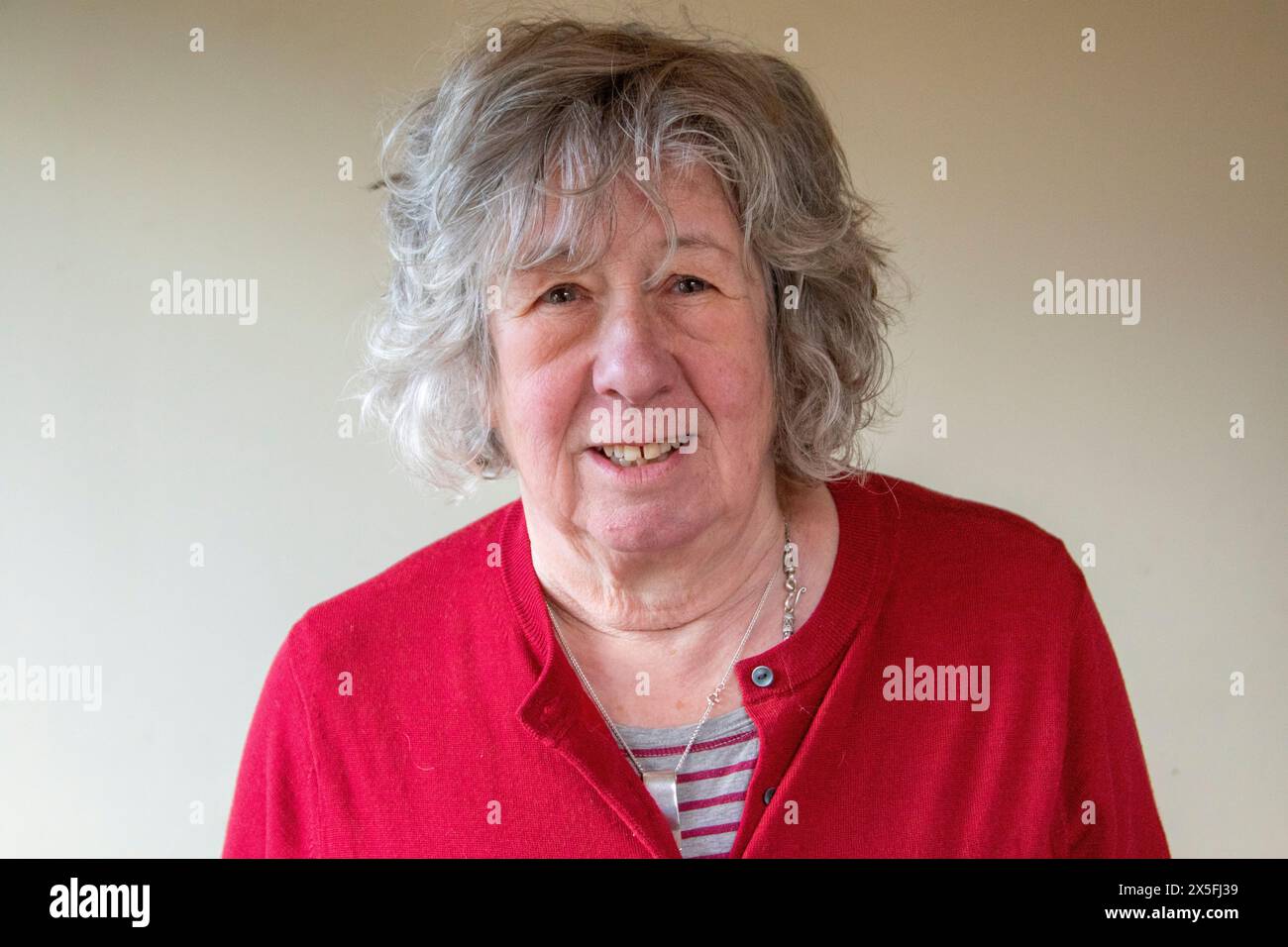 A mature woman with grey hair standing and in a dark red cardigan and mostly looking at the camera with a serious expression Stock Photo