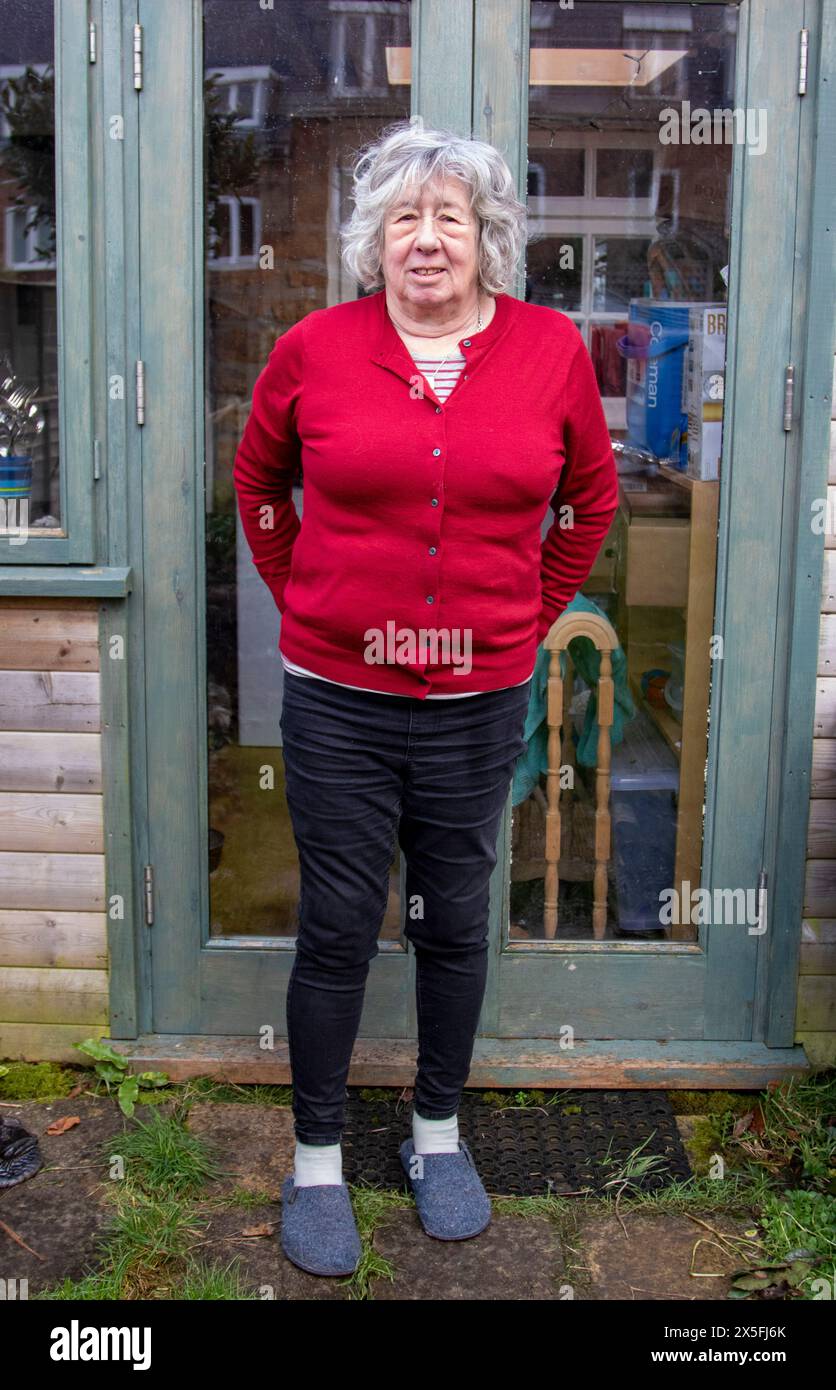 A mature woman with grey hair standing and in a dark red cardigan and mostly looking at the camera with a serious expression Stock Photo