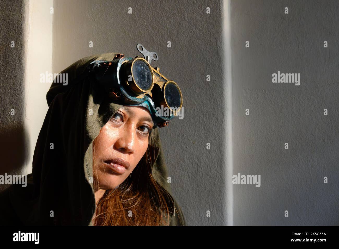 portrait of a woman donned in a hood and steampunk goggles, her gaze intensified by the striking contrast of sunlight and shadows Stock Photo