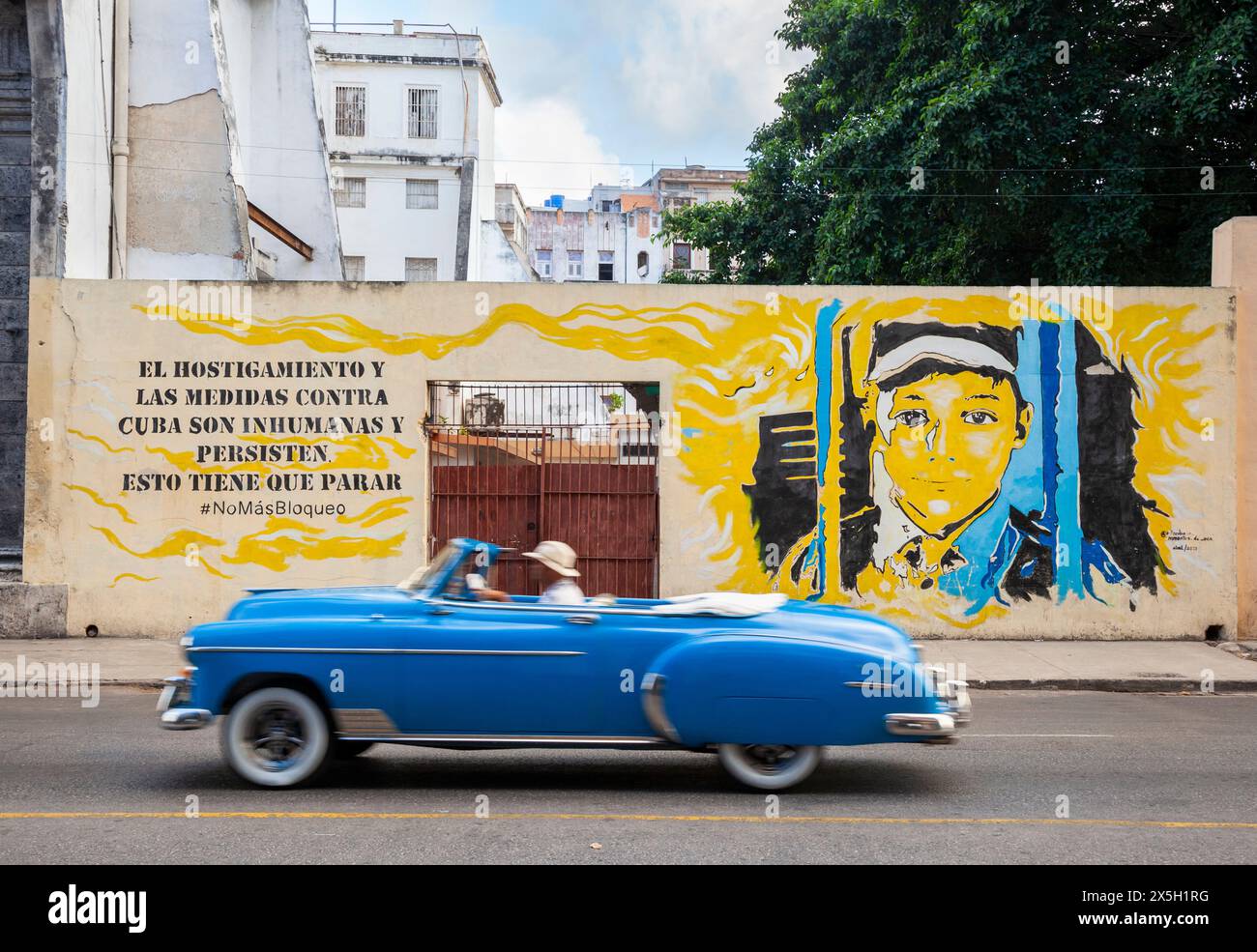 1950s Chevy passes Havana mural. The text reads: 'The harassment and measures against Cuba are inhuman and persist, this has to stop. No more blockade Stock Photo