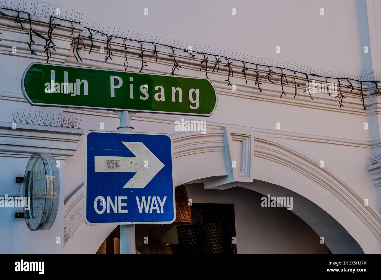 An image capturing a vibrant Jln Pisang street sign next to a blue one way directional arrow on a white architectural background with security feature Stock Photo