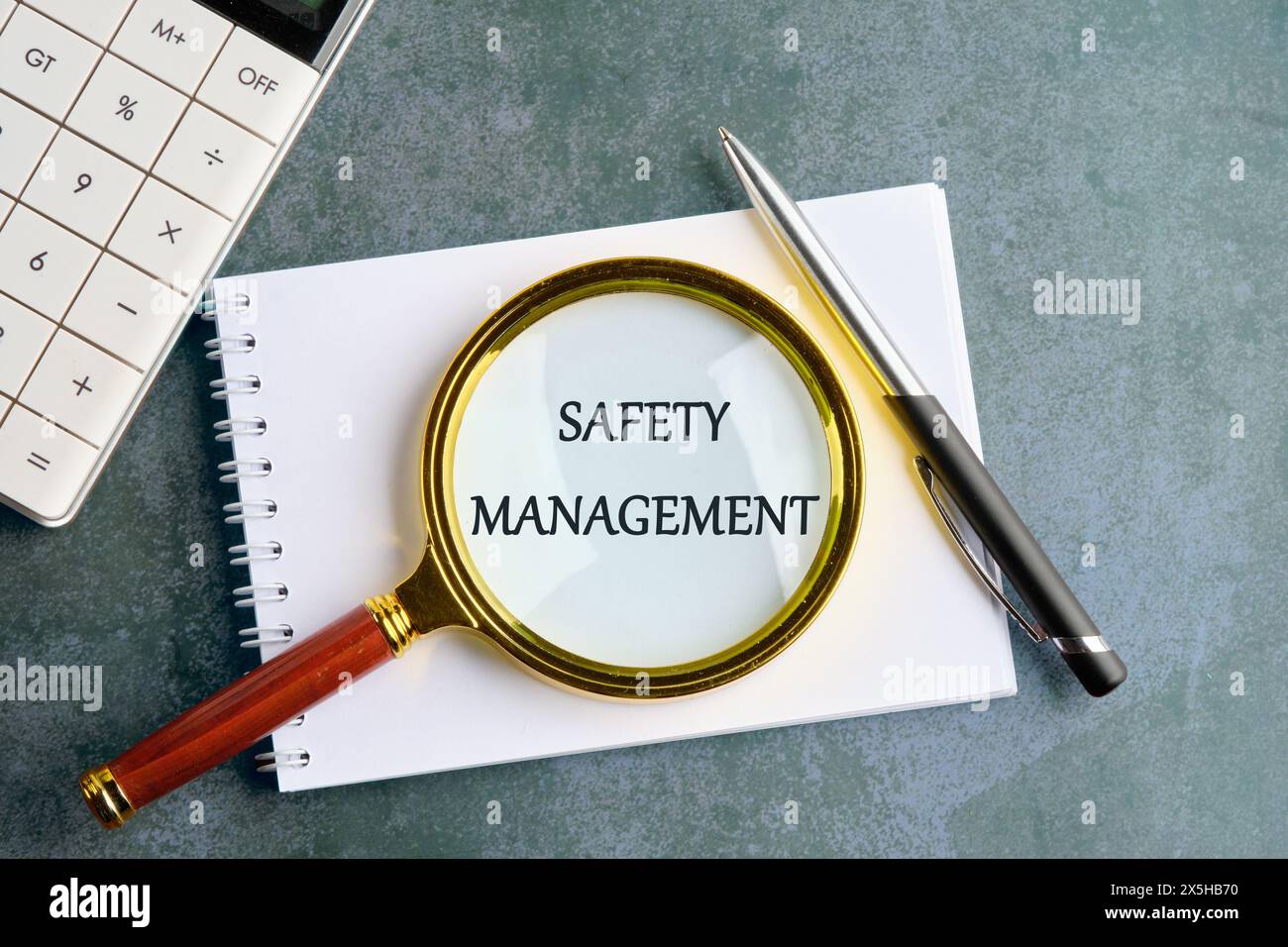 Safety Management text written through a magnifying glass on a notebook in a composition with a calculator, a pen on a gray background Stock Photo