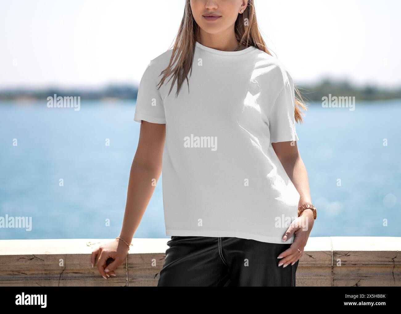 Mockup of a white T-shirt with a round neck on a girl, standing on the embankment, front view. Stylish female shirt template, woman posing on the back Stock Photo