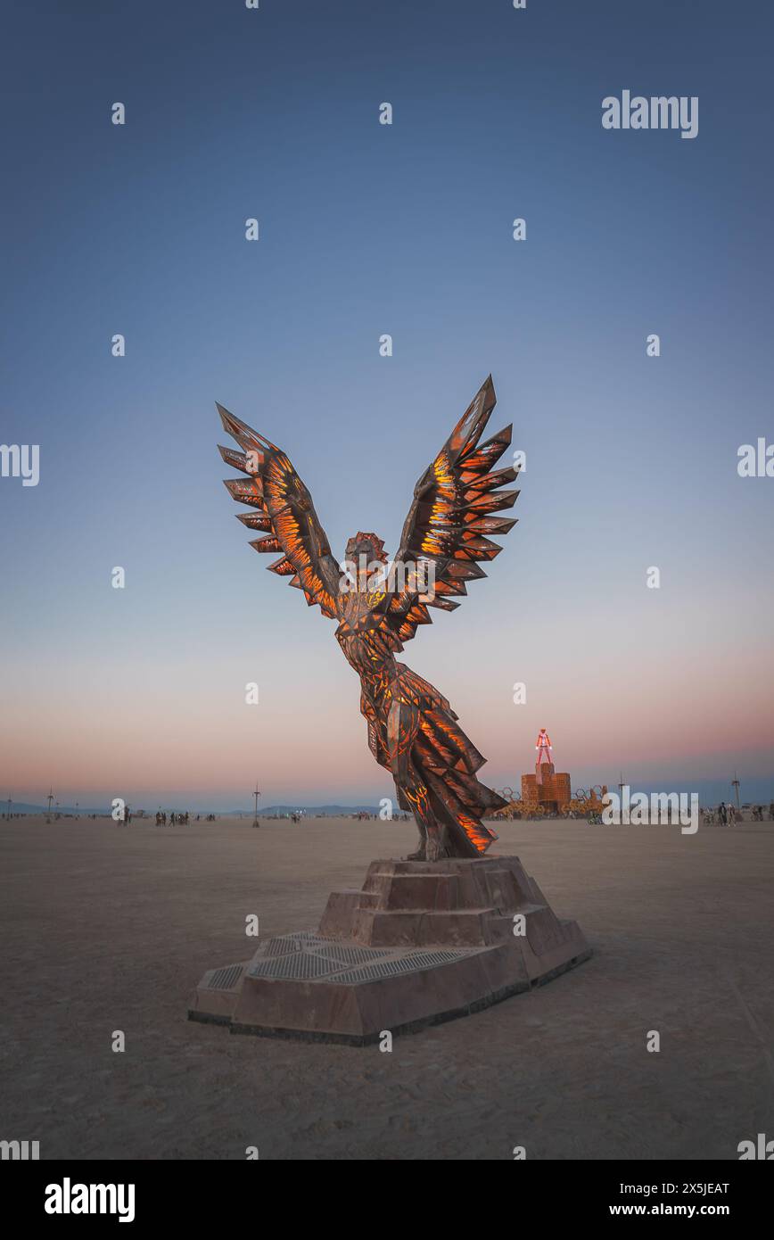 Angel sculpture with fiery wings against desert sky at dusk, art installation at music festival. Stock Photo