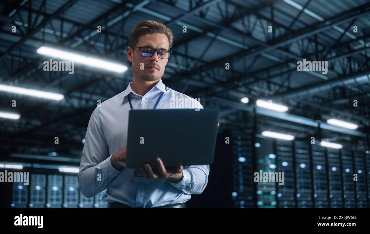 Male IT Engineer Working with Laptop in Data Center. Technician or Developer Standing at the Server Rack Corridor with a Laptop Computer. High-Speed Data Transfer, Analytics, Statistics Concept Stock Photo