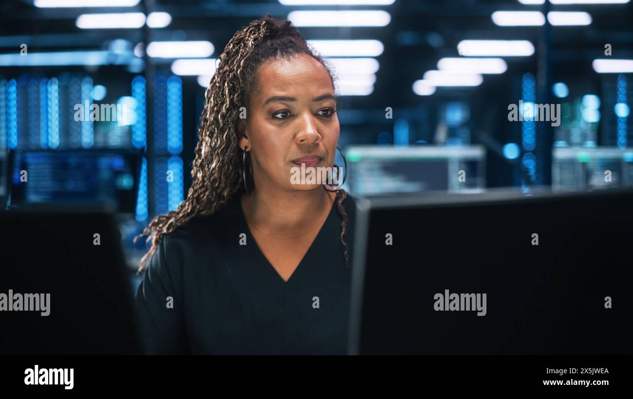 Focused IT Specialist Working on Computer in Office Environment. Diverse Multiethnic Female Developer is Using Two Monitors while Writing Code Late at the Night Stock Photo