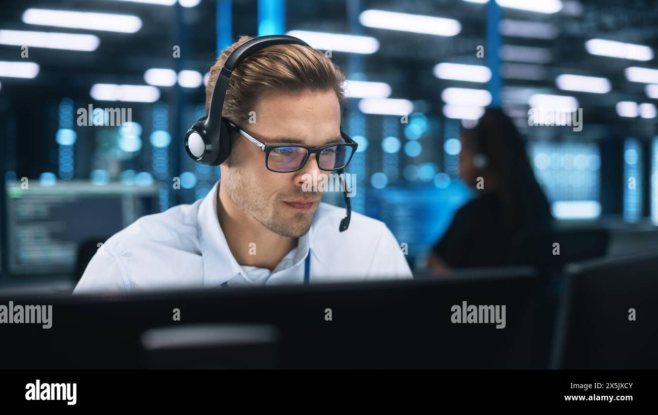 Busy Technical Customer Support Specialist is Talking on Headset while Working on a Computer in a Call Center Control Dark Room During the Evening. Customer Service Concept Stock Photo