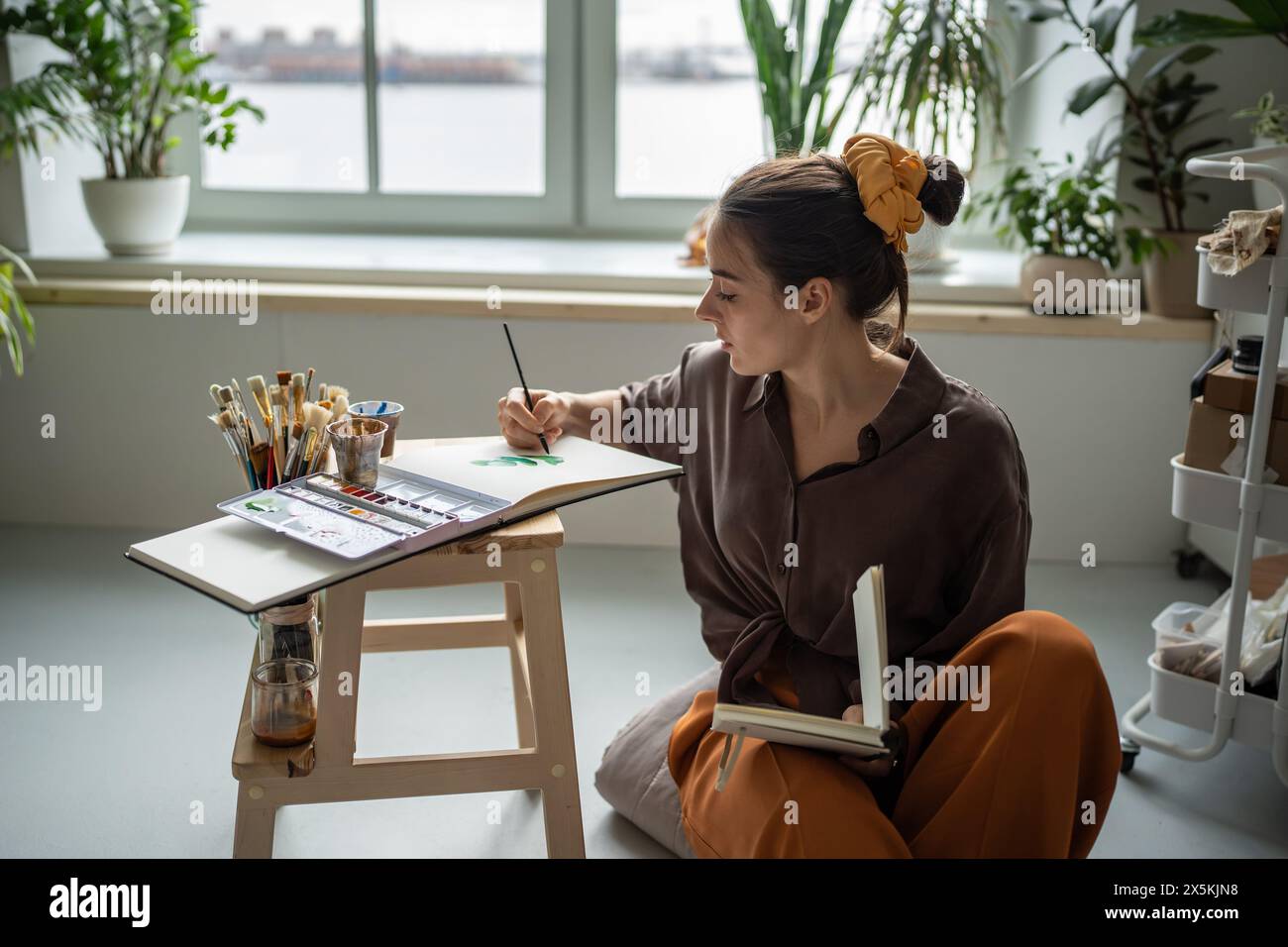 Unsmiling woman artist painting with watercolors. Female painter sitting at floor of home art studio Stock Photo