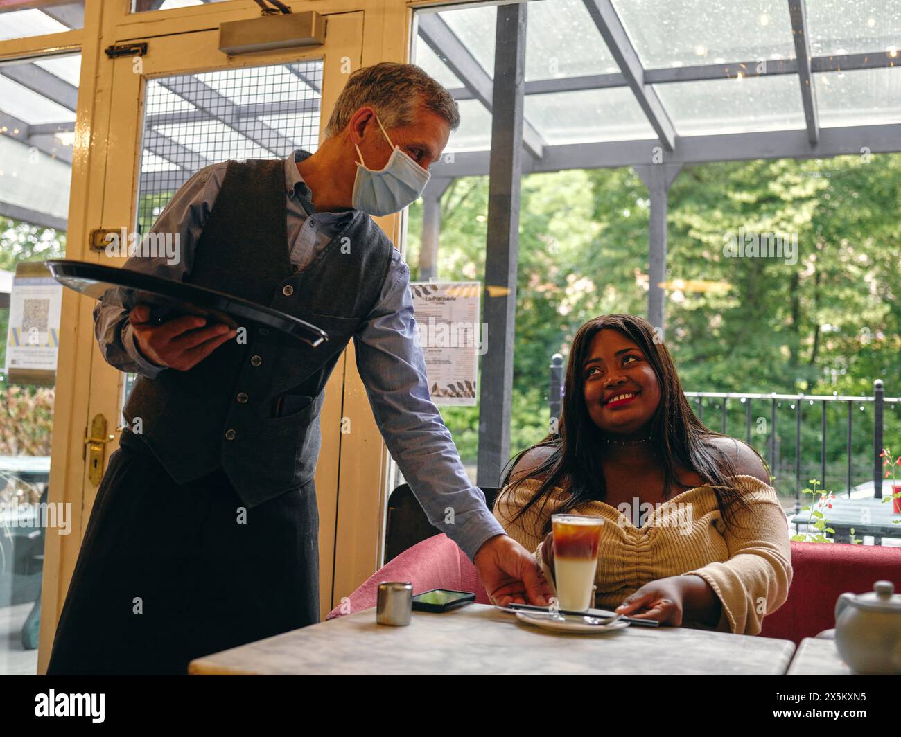 Waiter in face masks serving latte to smiling woman in cafe Stock Photo