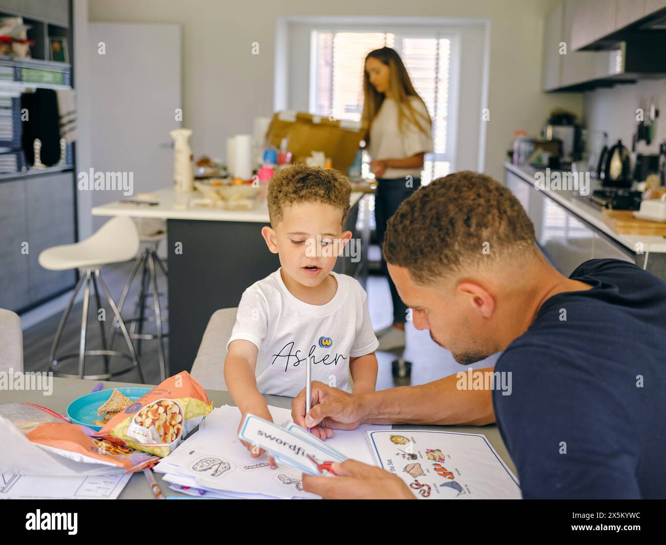 Father assisting son doing homework at home, mother in background Stock Photo