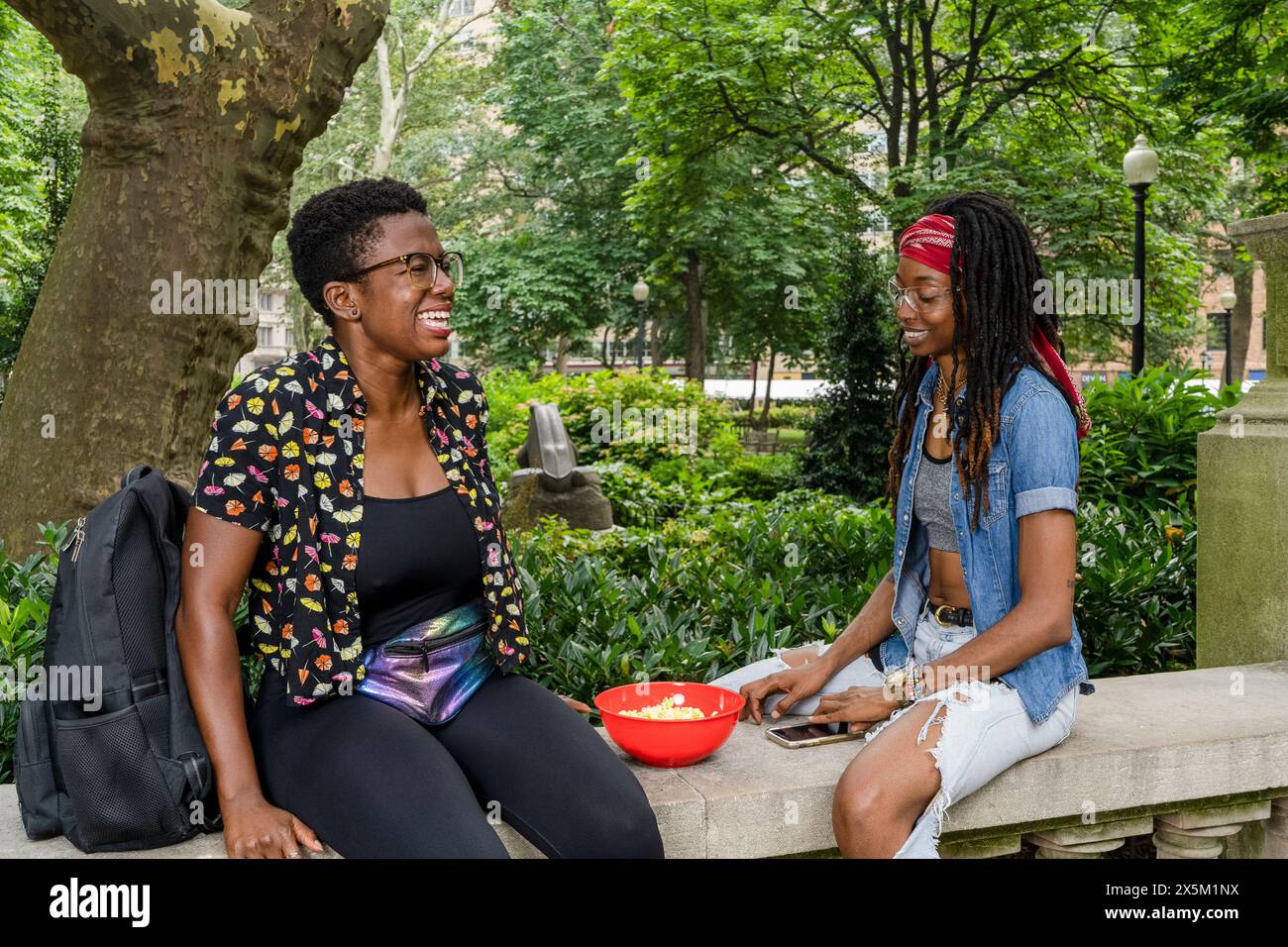 USA, Two smiling young women sitting on wall in park Stock Photo