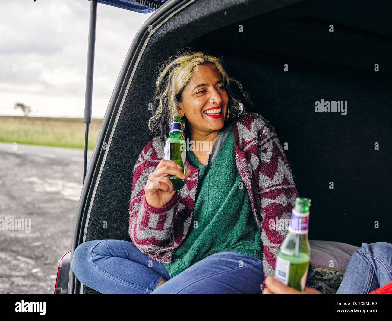 Woman sitting in back of van and holding beer Stock Photo