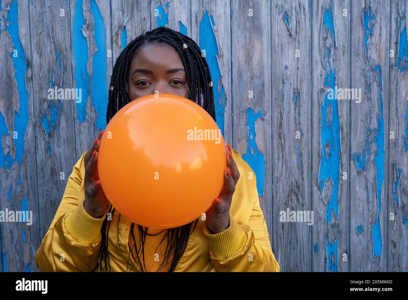 UK, South Yorkshire, Portrait of woman with braided hair blowing orange balloon Stock Photo