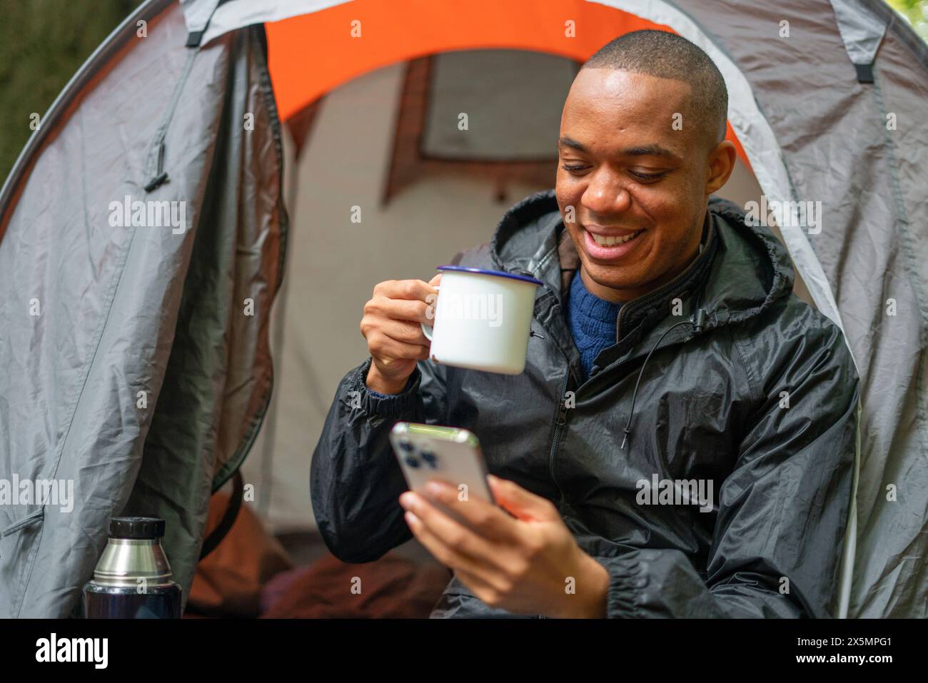 Smiling man using smart phone outside of tent and holding metal mug Stock Photo