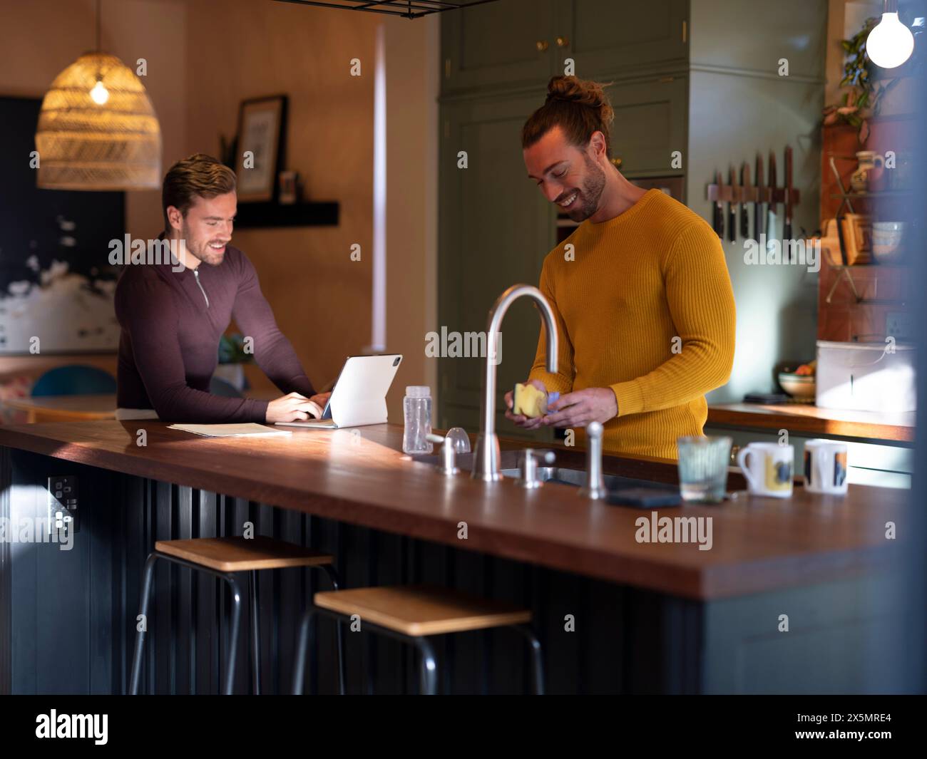 Man washing dishes while boyfriend using digital tablet at home kitchen Stock Photo