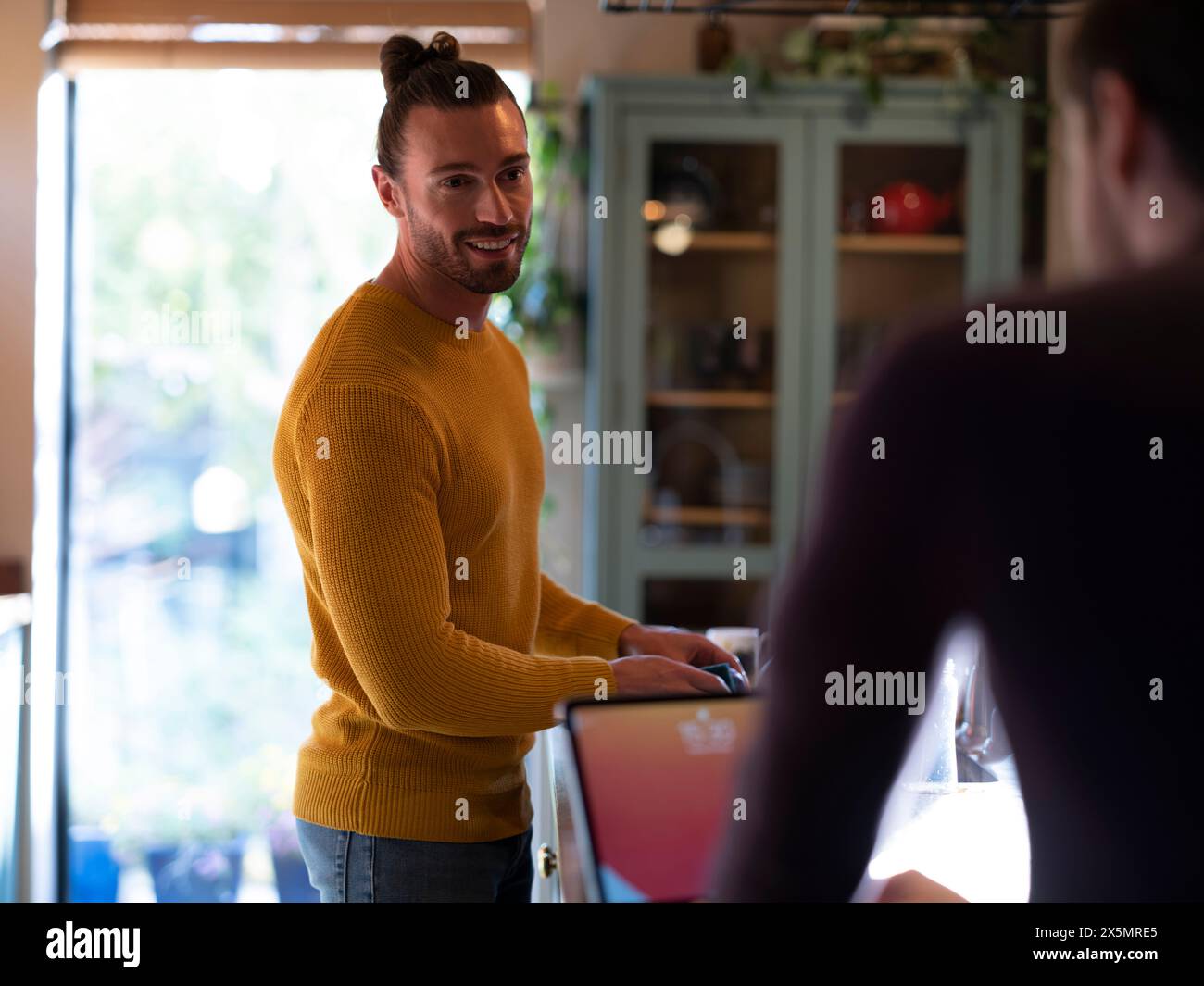 Man washing dishes while boyfriend using digital tablet at home kitchen Stock Photo