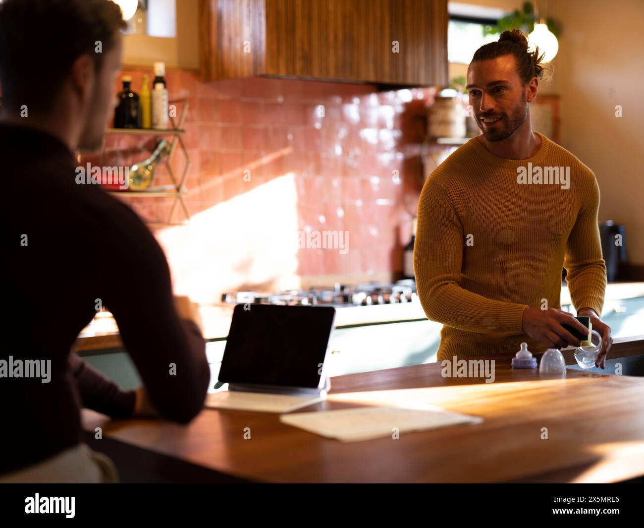 Man washing dishes while boyfriend using digital tablet at home kitchen Stock Photo