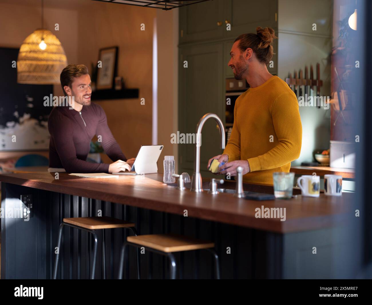 Man washing dishes while boyfriend using digital tablet at home kitchen Stock Photo