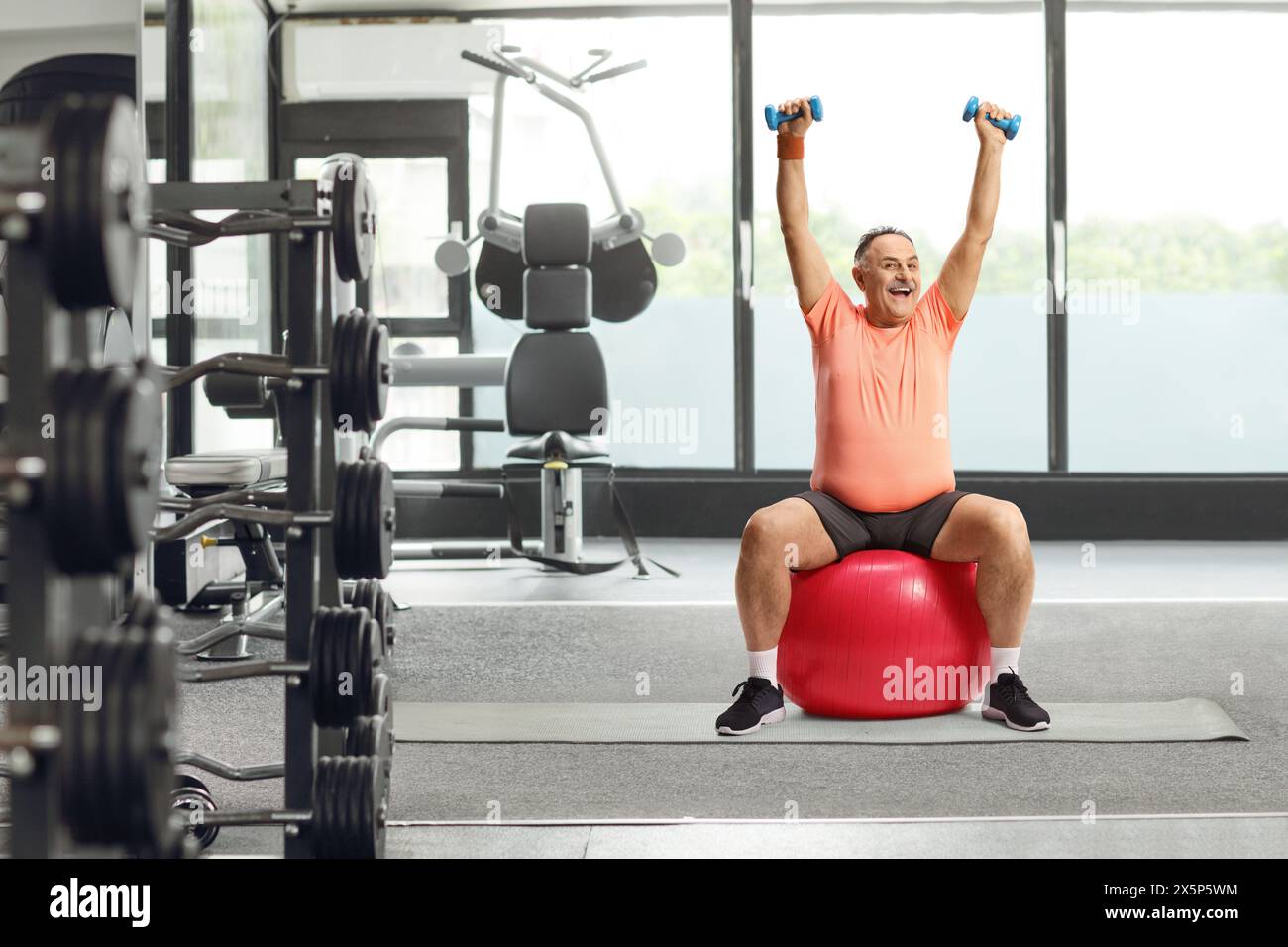 Smiling mature man in sportswear sitting on a fitness ball and exercising at a gym Stock Photo