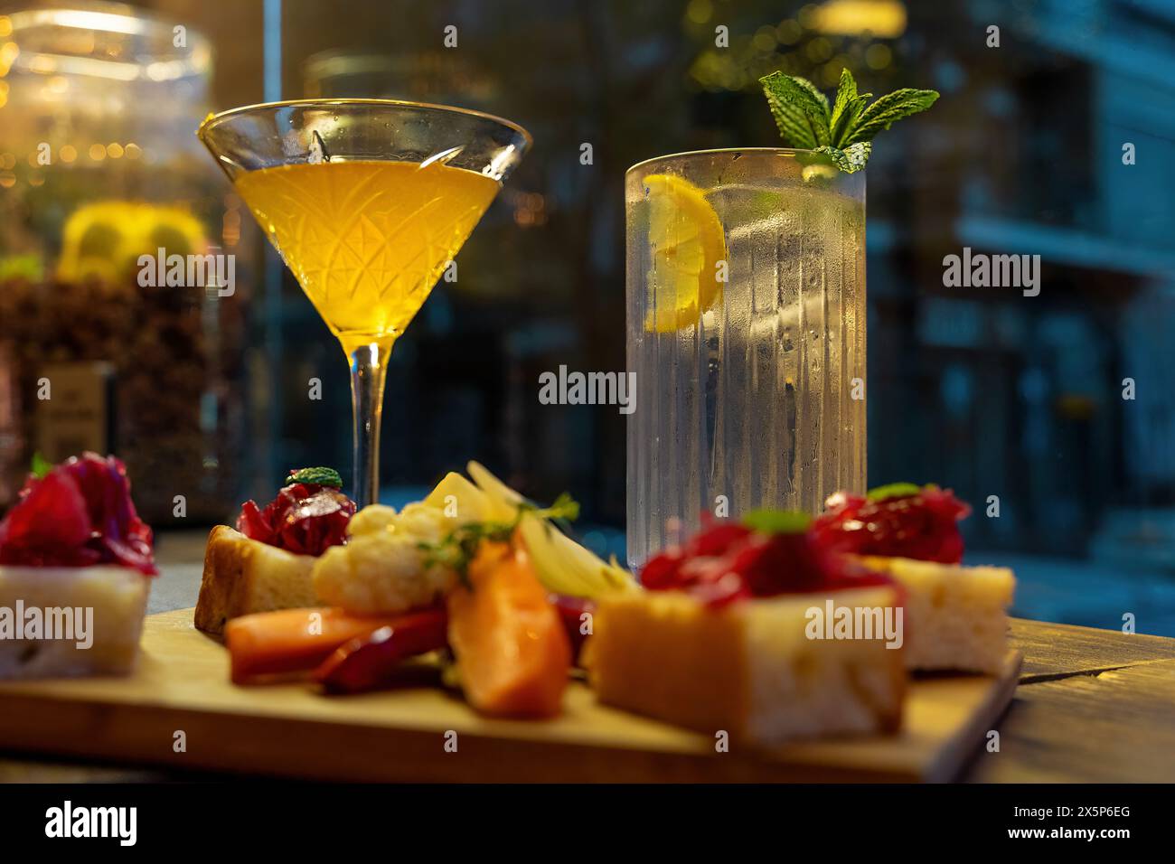 Cocktails and appetizers on a wooden board - gourmet bruschetta, refreshing drinks - evening indulgence - elegant glassware, vibrant garnish - dimly l Stock Photo