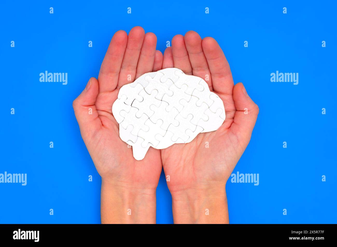 Hands holding a white puzzle piece in the shape of a brain against a vibrant blue background. Creativity, problem-solving and neuroscience related con Stock Photo