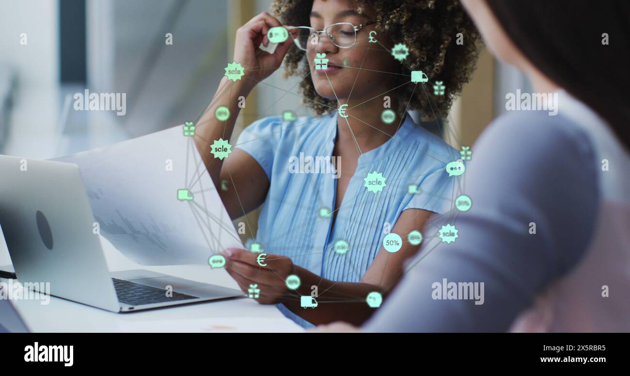 Image of globe of icons over two diverse female office colleagues discussing over a document Stock Photo