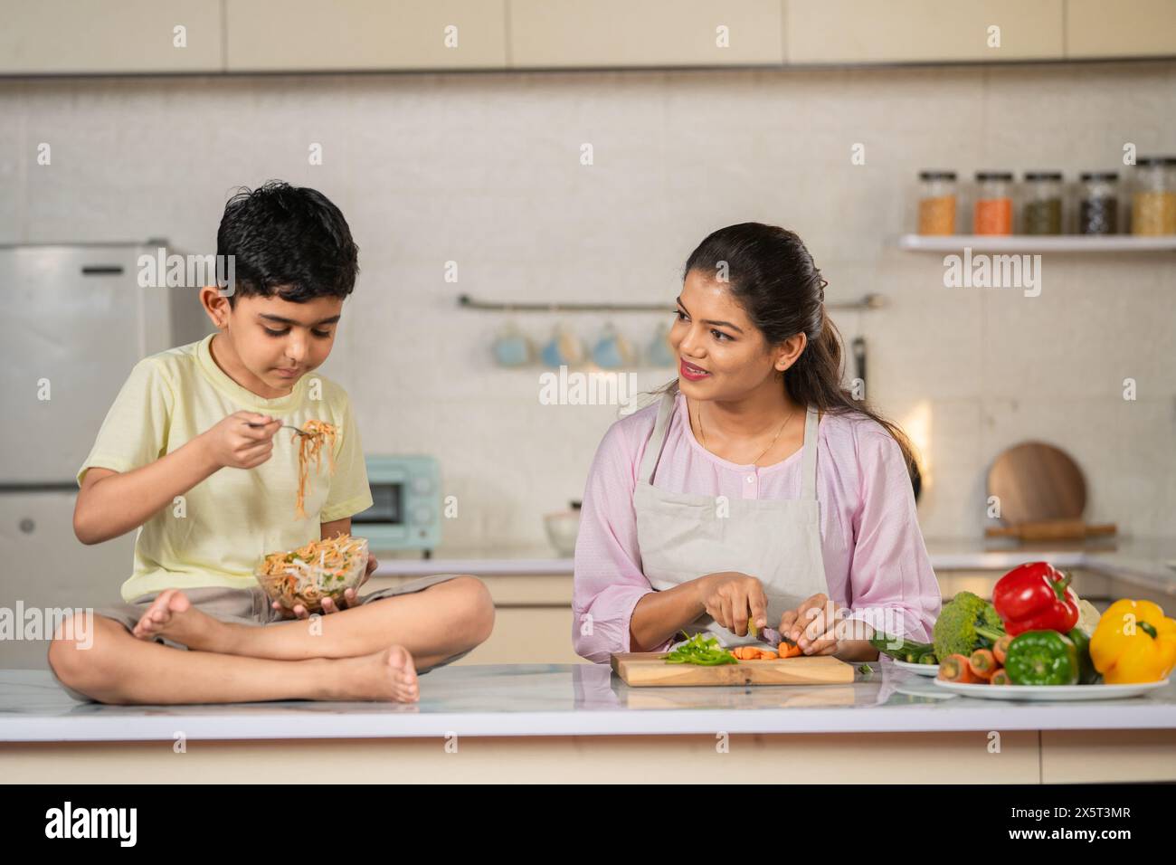 Joyful kid eating noodles at kitchen while spending time with working young mother kitchen - concept of childhood bonding, parenthood and healthy Stock Photo