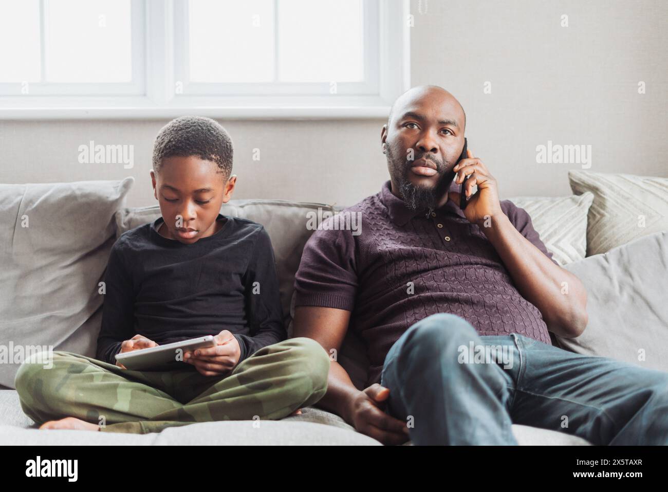 Father using phone sitting on sofas next to son (10-11) using digital tablet Stock Photo