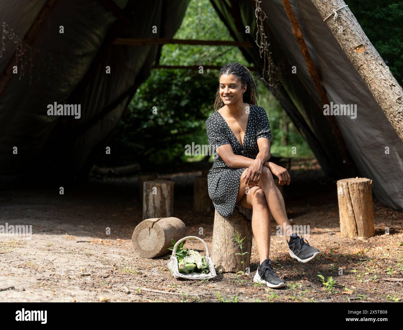 Smiling woman resting on tree stump in front of hut in forest Stock Photo