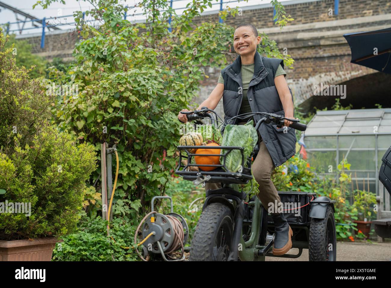 Smiling woman riding cargo electric trike loaded with homegrown vegetables Stock Photo