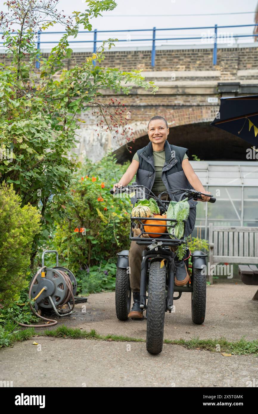 Smiling woman riding cargo electric trike loaded with homegrown vegetables Stock Photo