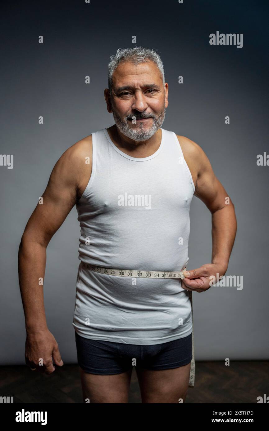 Portrait of man measuring waist against gray background Stock Photo