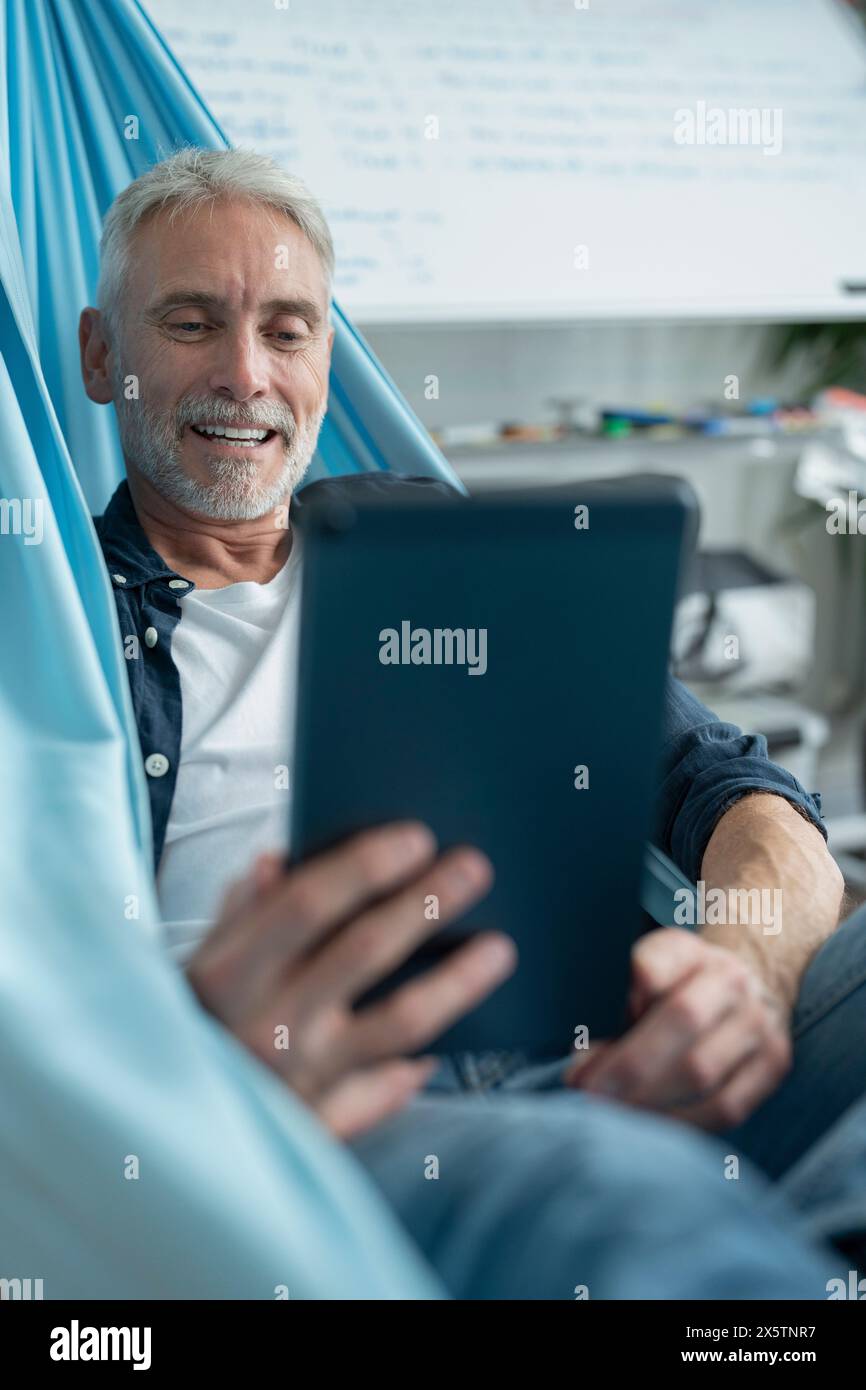Smiling mature man lying in hammock and using digital tablet Stock Photo