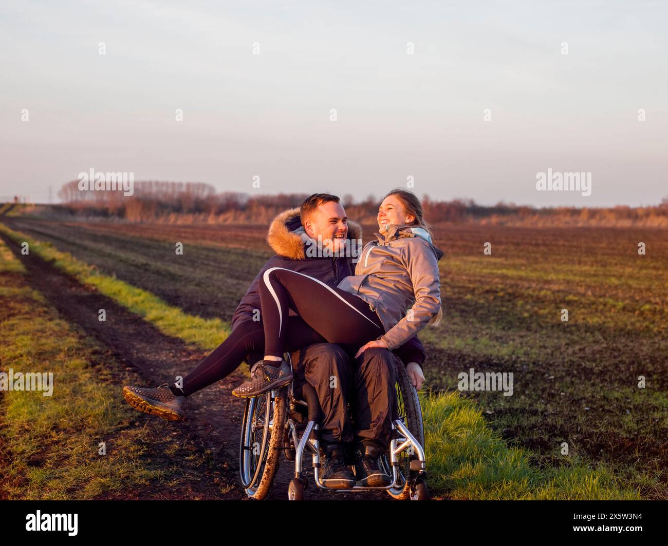 Smiling man on wheelchair in field with woman on laps Stock Photo